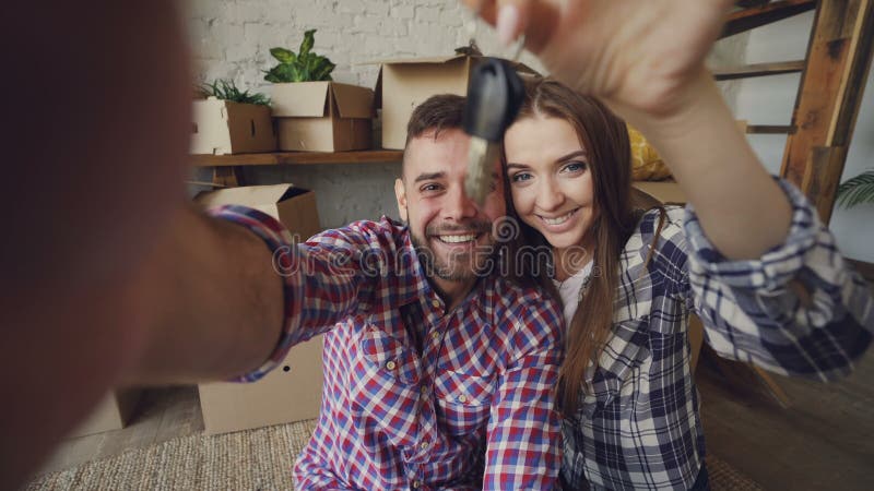 Point of view shot of happy couple taking selfie with house keys after purchasing new apartment. Young people are