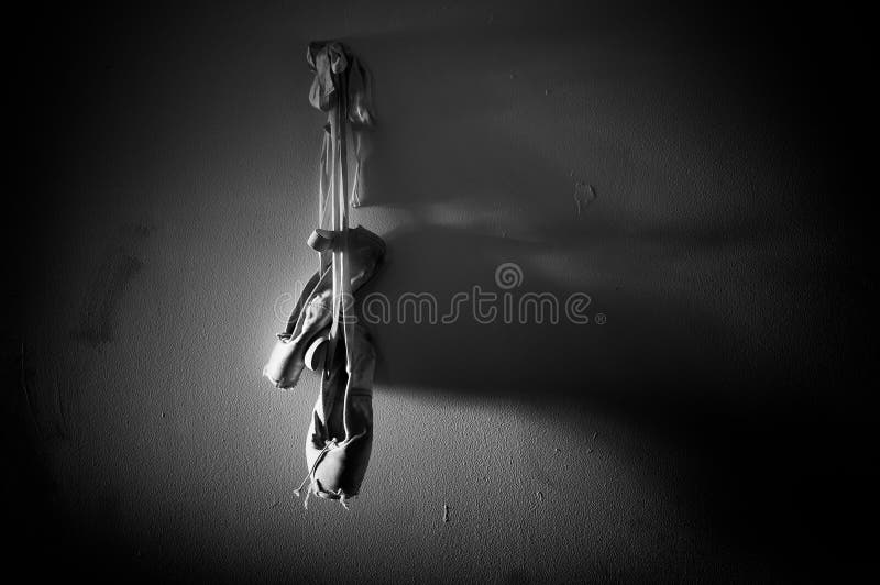 A worn out pair of ballet pointe sose is hanging from a wall with dramatic lighting and lit from the left in this black and white image with plenty of copy space. A worn out pair of ballet pointe sose is hanging from a wall with dramatic lighting and lit from the left in this black and white image with plenty of copy space.