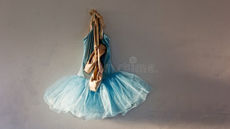 A blue velvet romantic tutu is hanging on a wall in dressing room with a worn pair of ballet pointe shoes on top of dress with copy or tesxt space, lit only by sunlight through window. A blue velvet romantic tutu is hanging on a wall in dressing room with a worn pair of ballet pointe shoes on top of dress with copy or tesxt space, lit only by sunlight through window