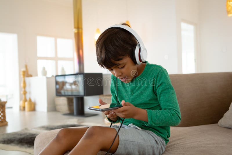 Pojke med hörlurar med mikrofon som spelar leken på mobiltelefonen på soffan i ett bekvämt hem royaltyfria foton Pojke med hörlurar med mikrofon som spelar leken på mobiltelefonen på soffan i ett bekvämt hem royaltyfria foton
