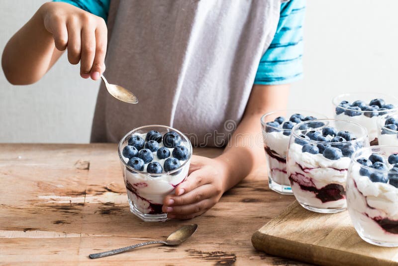 Pojken rymmer den hemlagade efterrätten med grekisk yoghurt eller kräm, blåbärdriftstopp och nya blåbär royaltyfri foto Pojken rymmer den hemlagade efterrätten med grekisk yoghurt eller kräm, blåbärdriftstopp och nya blåbär royaltyfri foto