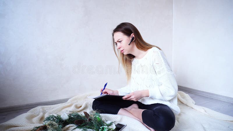 Sweet female professional photographer talks to customer and specifies details of New Year`s photography, writes pen in black folder with documents. girl with good mood smiles and laughs, sits on floor on knitted plaid near Christmas pine wreath and flickering garland in bright room in daytime. European-looking Woman with long blond hair is dressed in white sweater and black pants. Concept of gadgets and modern technology, consumption and training of new devices, remote work and entrepreneurship, festive mood and New Year or Christmas Eve, happy and beautiful people, holiday attributes and symbols. Sweet female professional photographer talks to customer and specifies details of New Year`s photography, writes pen in black folder with documents. girl with good mood smiles and laughs, sits on floor on knitted plaid near Christmas pine wreath and flickering garland in bright room in daytime. European-looking Woman with long blond hair is dressed in white sweater and black pants. Concept of gadgets and modern technology, consumption and training of new devices, remote work and entrepreneurship, festive mood and New Year or Christmas Eve, happy and beautiful people, holiday attributes and symbols.