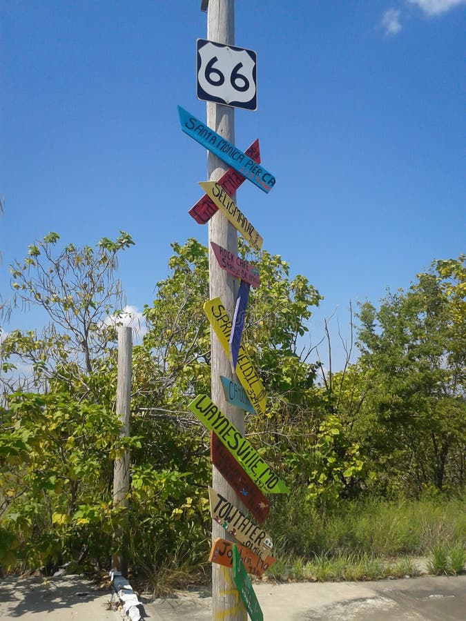 Pole with Route 66 highway shield at top and place names below on quirky colored signs along old stretch of highway. Pole with Route 66 highway shield at top and place names below on quirky colored signs along old stretch of highway