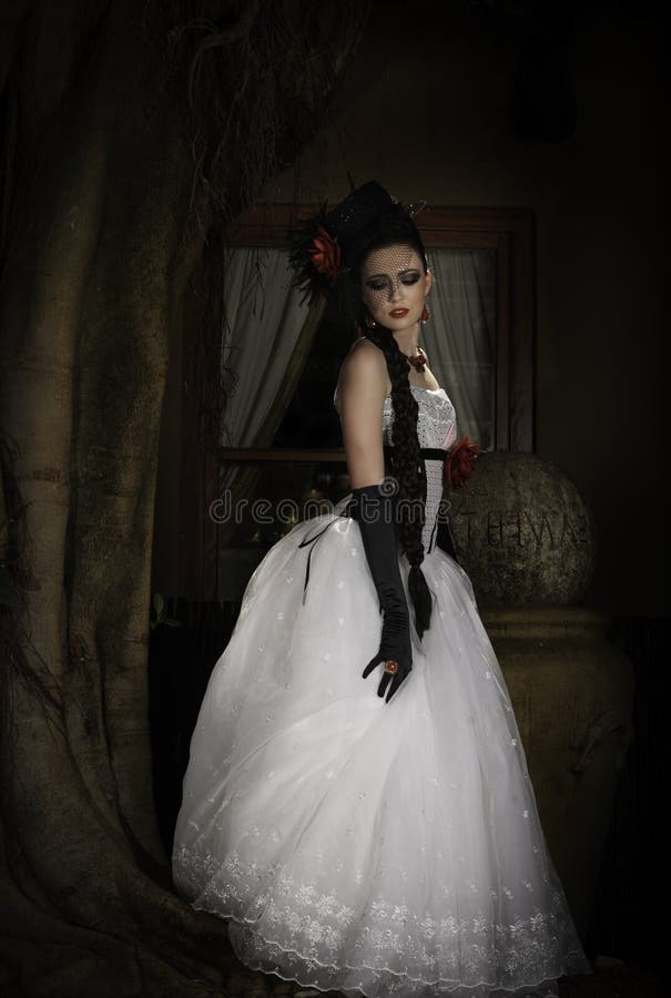 Portrait of a beautiful young woman in white and black wedding dress with embroidered lace skirt and red fabric flower detail, wearing long gloves, a hat with net covering her face, earrings and necklace, with her long dark hair styled in an elaborate braid, posing outside a window next to a tree. Portrait of a beautiful young woman in white and black wedding dress with embroidered lace skirt and red fabric flower detail, wearing long gloves, a hat with net covering her face, earrings and necklace, with her long dark hair styled in an elaborate braid, posing outside a window next to a tree.