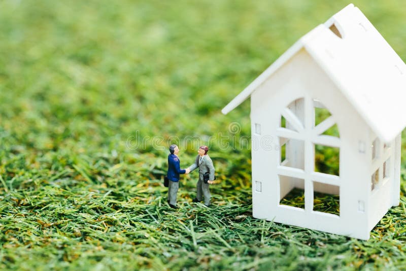Popolo in miniatura, uomo d'affari di successo in piedi e stretta di mano con una casa bianca di legno sull'erba verde Investimen fotografie stock libere da diritti Popolo in miniatura, uomo d'affari di successo in piedi e stretta di mano con una casa bianca di legno sull'erba verde Investimen fotografie stock libere da diritti