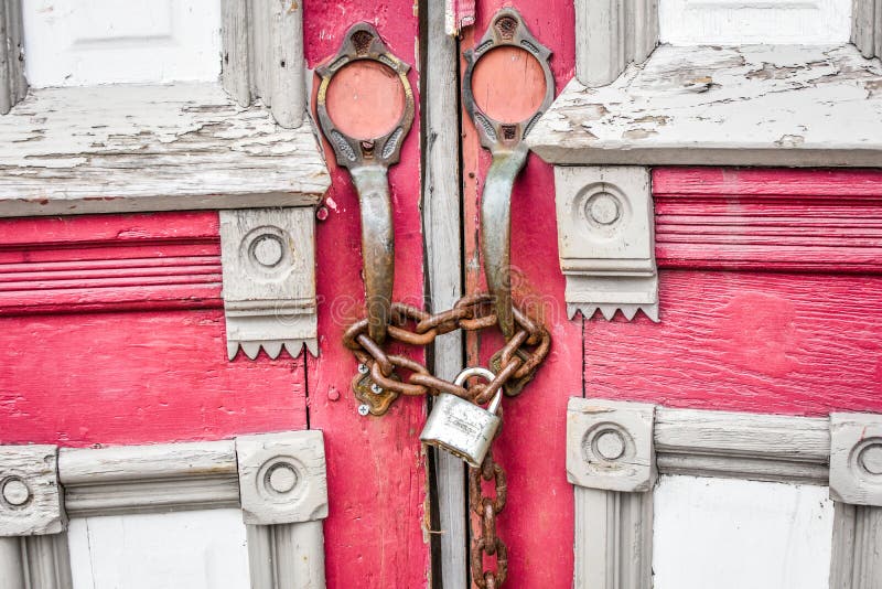 A close up of a set of doors padlocked together with a rusty chain. The doors were from an abandoned church building. The doors are red and gray with pink, antique handles. The door has ornate decoration and weathered wood. A close up of a set of doors padlocked together with a rusty chain. The doors were from an abandoned church building. The doors are red and gray with pink, antique handles. The door has ornate decoration and weathered wood.
