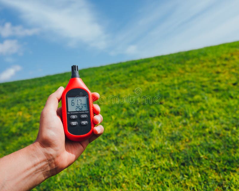 Portable thermometer in hand measuring outdoor air temperature and humidity, green field and blue sky background. Portable thermometer in hand measuring outdoor air temperature and humidity, green field and blue sky background