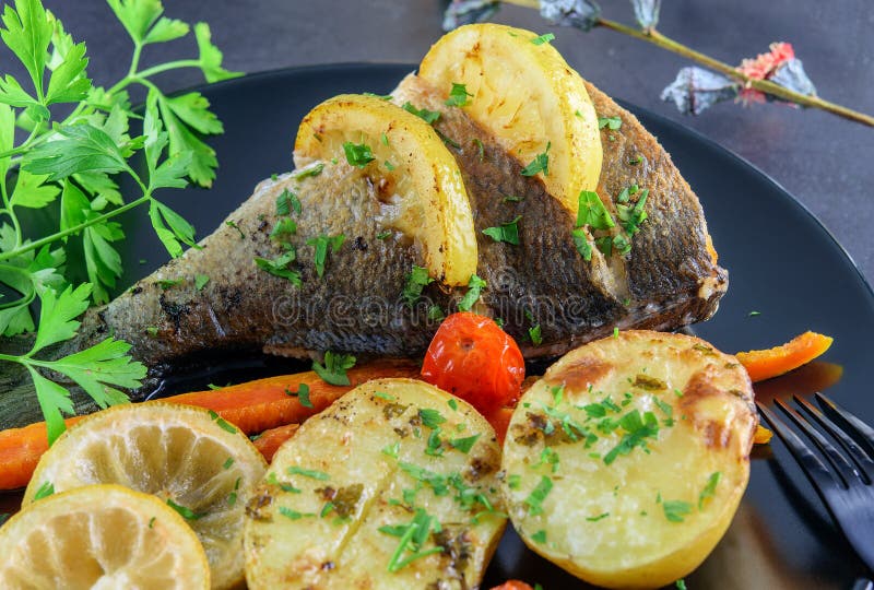 Fried fish dorada on the black dish and on the black background with lemon and some vegetables closeup. Healthy mediterranean food stock photo