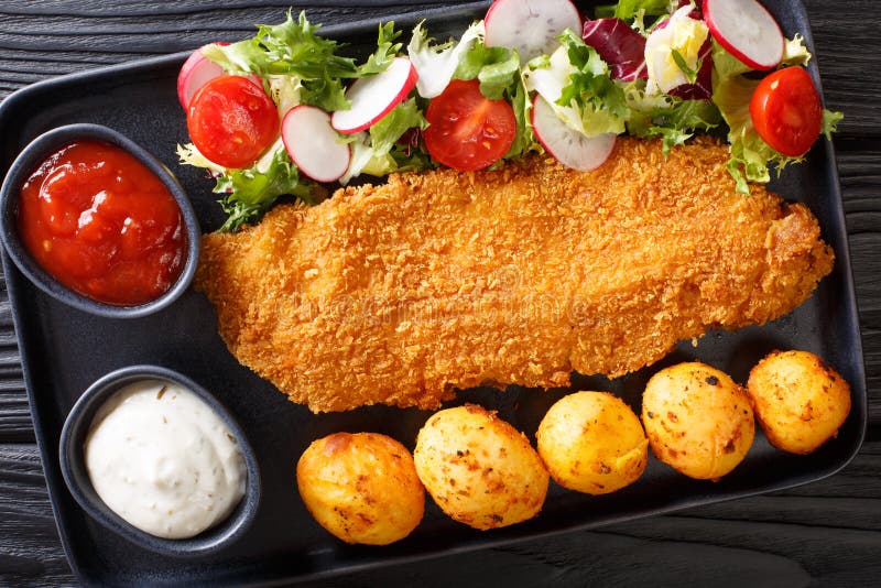 Portion of white fish fillet in breading with new potatoes and fresh vegetable salad close-up on the table. Horizontal top view