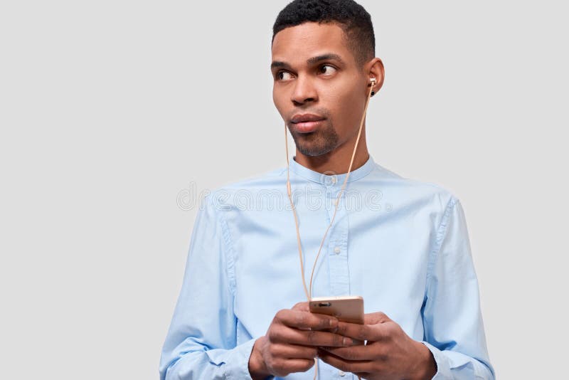 African American young man holds modern smart phone, blogging on cellphone. Handsome male listening the music on earphones stock photo African American young man holds modern smart phone, blogging on cellphone. Handsome male listening the music on earphones stock photo
