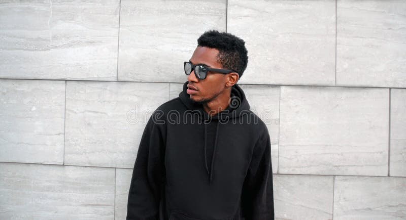 Portrait african man in black hoodie, sunglasses looking away on city street over gray brick wall