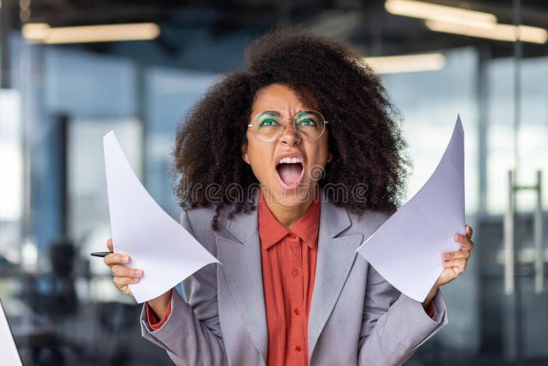 Portrait of angry african american woman holding documents and screaming while looking up in office. Crazy female in