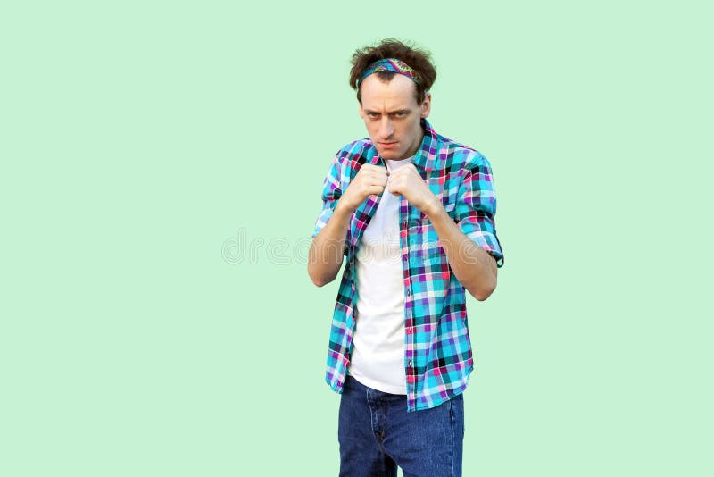 Portrait of angry young man in casual blue checkered shirt and headband standing and looking at camera with boxing fists and ready stock photos Portrait of angry young man in casual blue checkered shirt and headband standing and looking at camera with boxing fists and ready stock photos