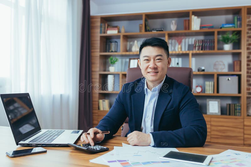 Portrait of Asian businessman working with papers and documents, man smiling and looking at camera, boss at work at desk in office