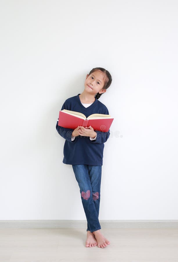 Portrait asian little child girl in casual school uniform reading a book and standing against white wall in the room