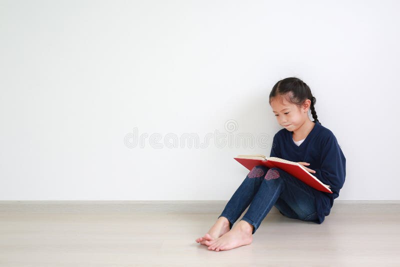 Portrait asian little kid girl in school uniform reading a book and sitting against white wall in the room with copy space