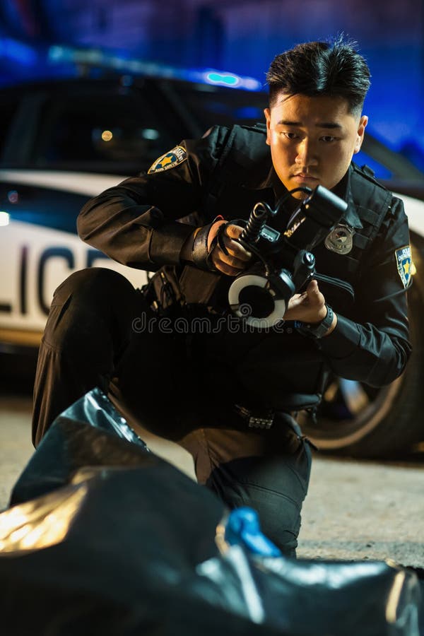 Portrait of Asian Police Officer Taking Forensics Photos of the Victim& x27;s Dead Corpse in a Body Bag on the Crime Scene. Young Detective from Police Department Solving his First Murder Case