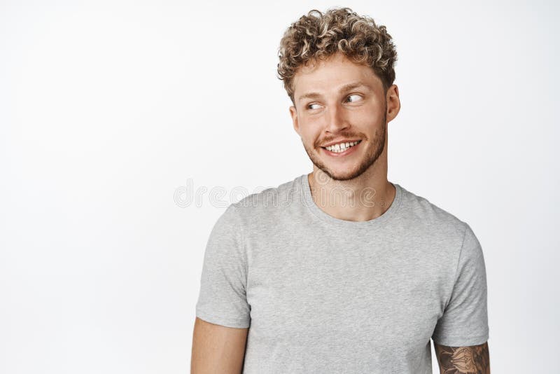 Portrait of attractive blond guy smiling, looking aside at copy space, wearing grey t-shirt, standing against white