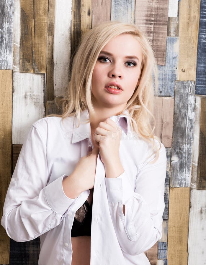 Portrait of attractive blonde girl standing on wood wall background. She has blue eyes and dressed in a man`s shirt