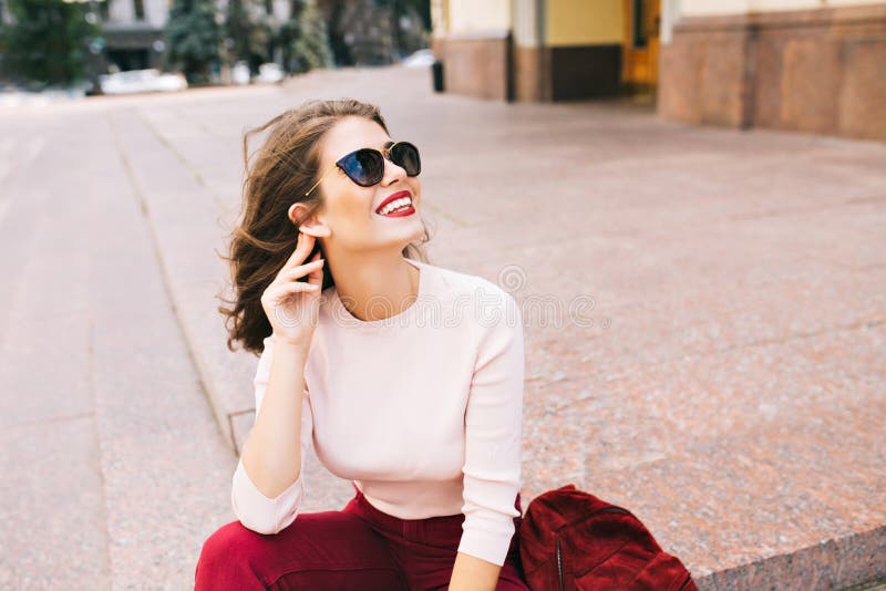 Portrait of attractive girl with snow-white smile and vinous lips sitting on stairs in city. She wears sunglasses an