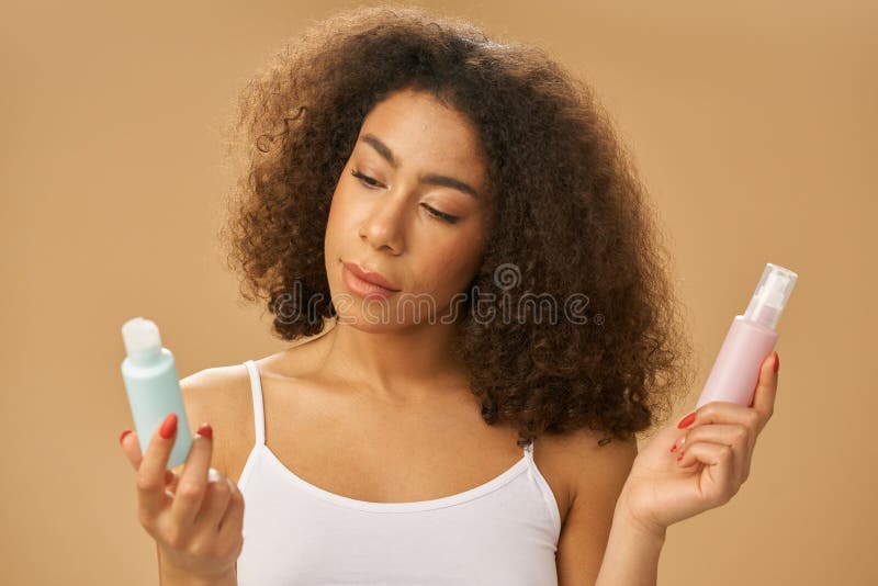 Portrait of attractive healthy african american young woman choosing between two bottles with beauty product while