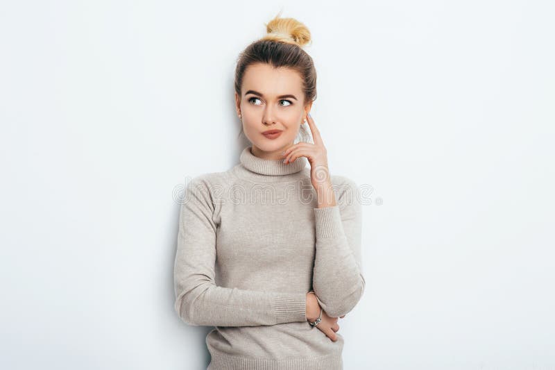 Portrait of attractive thoughtful young female wearing her blonde hair in knot holding forefinger up looking away with sly smile. Pretty girl having thoughtful dreamy look posing at studio wall