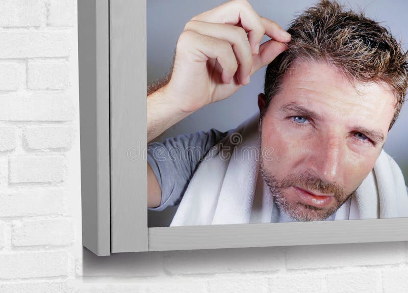 Portrait of attractive worried and concerned Caucasian man looking at bathroom mirror finding himself with gray hair in head feeli