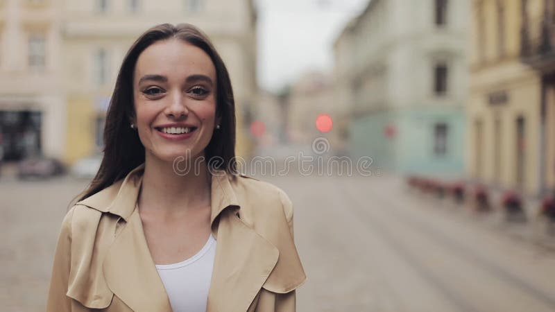 Portrait of Attractive Young Caucasian Girl Wearing Beige Trench Smiling and Laughing Standing at City Background.