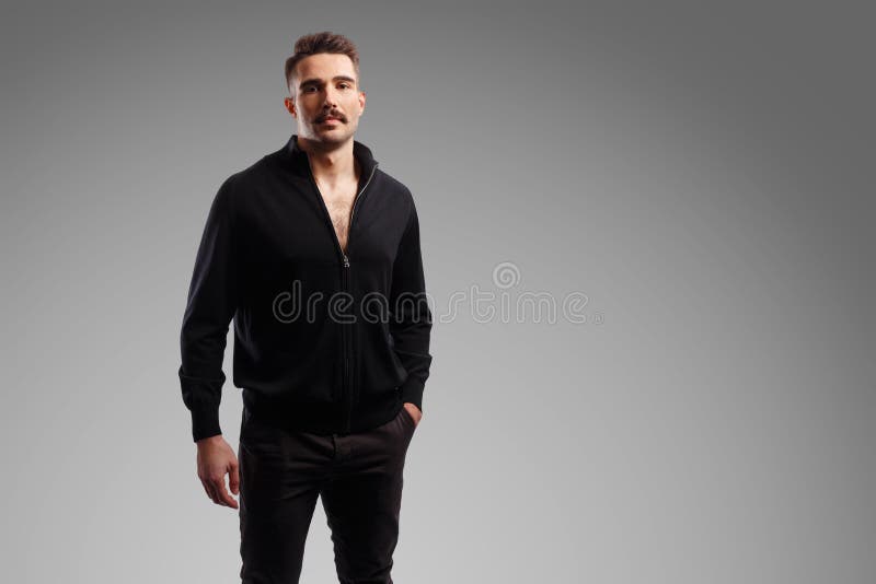 Portrait of a bearded bruntal man in black jacket, with a hand in his pocket, looking at camera over grey background. stock photo Portrait of a bearded bruntal man in black jacket, with a hand in his pocket, looking at camera over grey background. stock photo