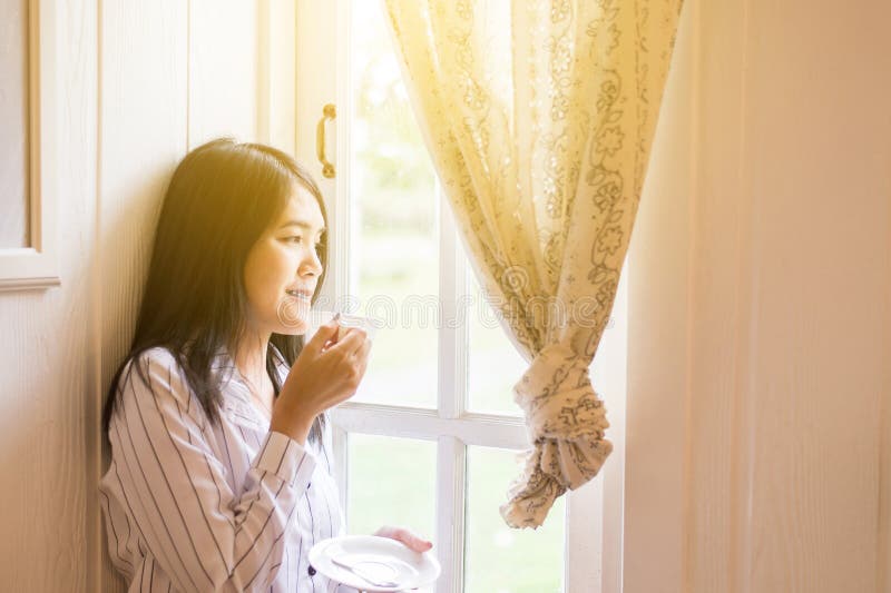 Portrait of beautiful asian woman is holding a cup of coffee and looking something on window at home in the morning,Happy and smil royalty free stock photos Portrait of beautiful asian woman is holding a cup of coffee and looking something on window at home in the morning,Happy and smil royalty free stock photos