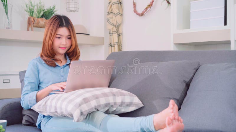 Portrait of beautiful attractive Asian woman using computer or laptop holding a warm cup of coffee or tea while lying on the sofa.