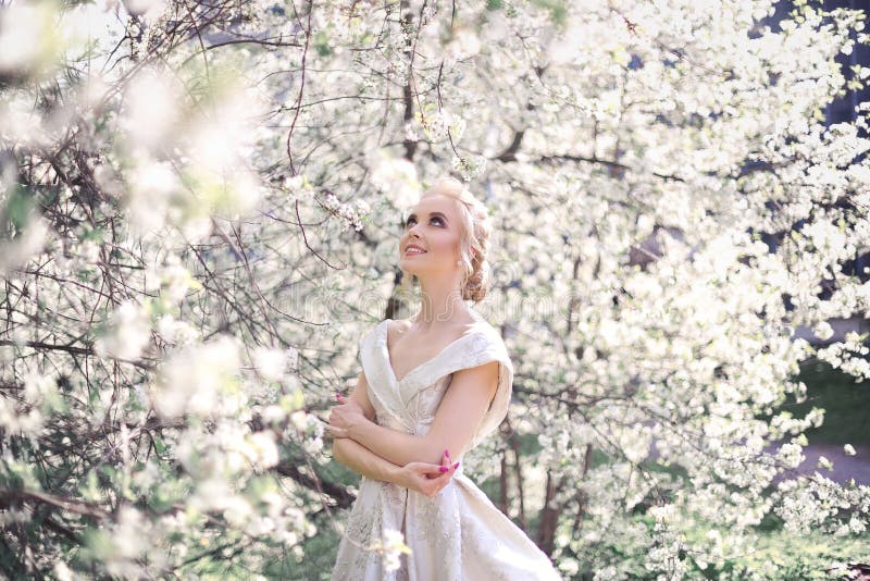 Portrait of a beautiful blonde girl in a luxurious white dress with open shoulders on the background of a blooming orchard.