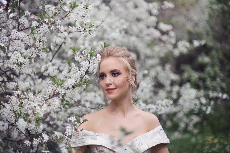 Portrait of a beautiful blonde girl in a luxurious white dress with open shoulders on the background of a blooming orchard.