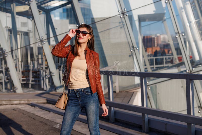 Portrait of beautiful brunette young woman in nice red brown jacket, denim jeans and sunglasses. Genuine leather bag, high heels royalty free stock photo Portrait of beautiful brunette young woman in nice red brown jacket, denim jeans and sunglasses. Genuine leather bag, high heels royalty free stock photo