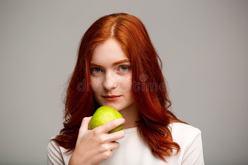 Portrait of beautiful ginger girl holding apple over gray background.