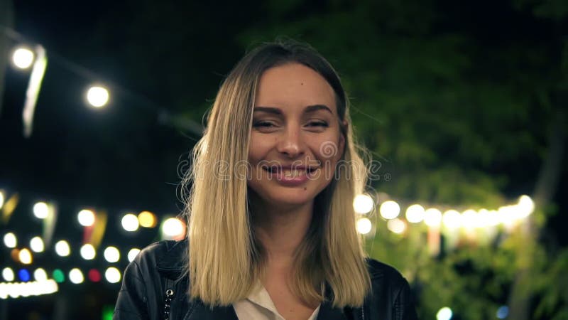 Portrait of a beautiful girl in a black leather jacket and white blouse standing in the night park. Light of a cafe