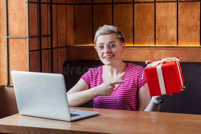 Portrait of beautiful happy young woman with short hair in pink t-shirt is sitting in cafe, pointing finger to red present box, stock photo Portrait of beautiful happy young woman with short hair in pink t-shirt is sitting in cafe, pointing finger to red present box, stock photo