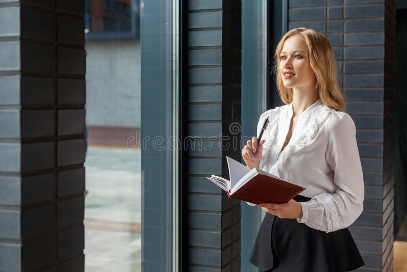 Portrait of beautiful inspired young woman making notes in diary book, looking out window while pondering idea. indoors