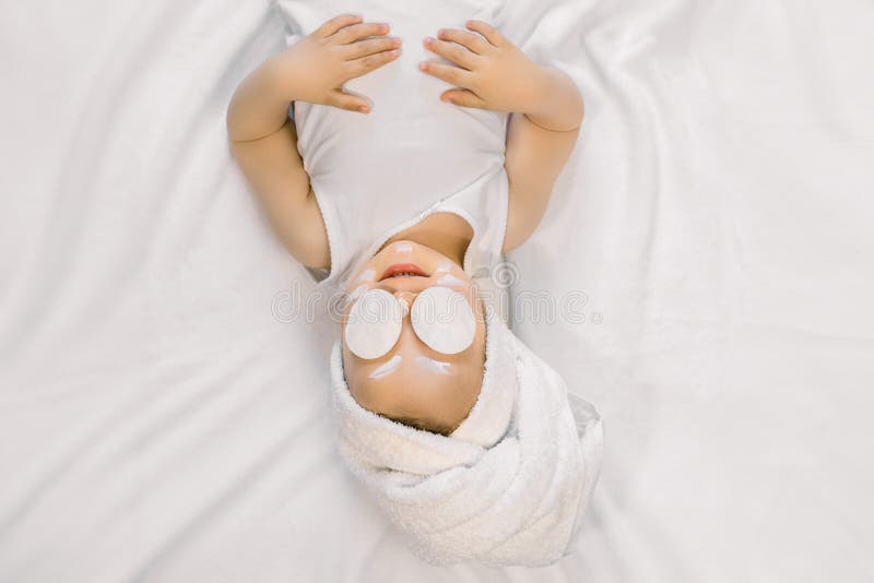 Portrait of beautiful little baby girl with white towel on her head, face cream on cheeks and forehead, and cotton pads