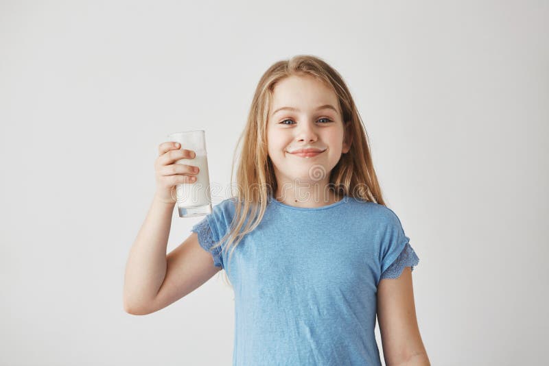 Portrait of beautiful small girl with blond long hair and blue eyes, smiling brightfully, holding glass of milk in hand