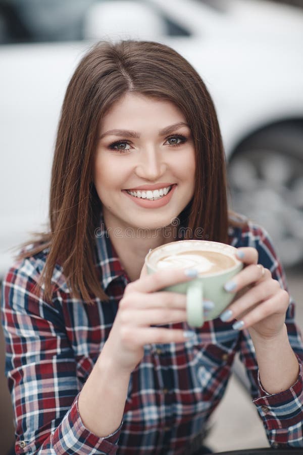 Pretty girl, beautiful brunette with elegant hairstyle in a plaid shirt drinks cup of coffee outdoors in summer cafe