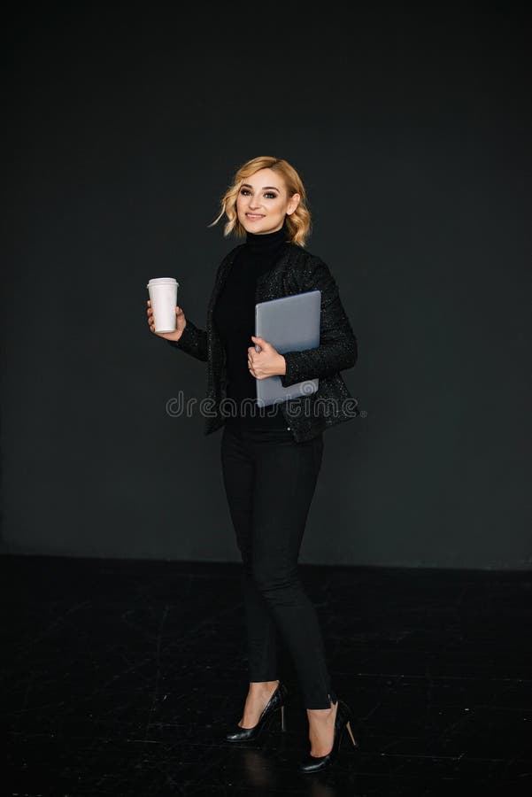 Portrait of a beautiful stylish businesswoman with a laptop and a glass of coffee in her hands in full growth on a black royalty free stock photography Portrait of a beautiful stylish businesswoman with a laptop and a glass of coffee in her hands in full growth on a black royalty free stock photography