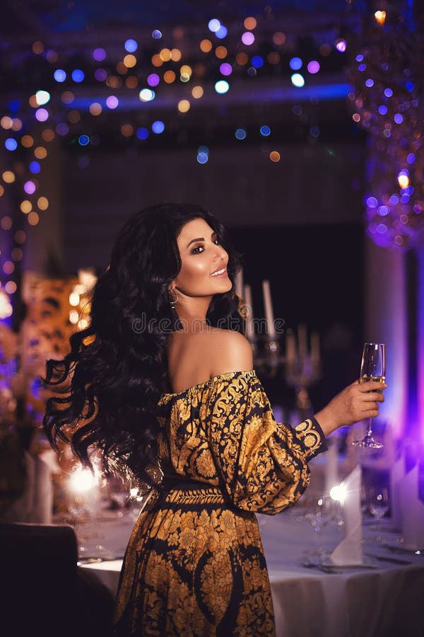 Portrait of a beautiful tanned brunette girl in a silk black and gold dress with a glass of champagne