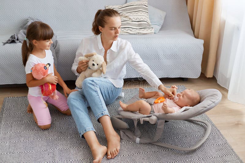 Portrait of beautiful woman wearing white shirt and jeans sitting on floor near sofa and playing with her infant baby and elder