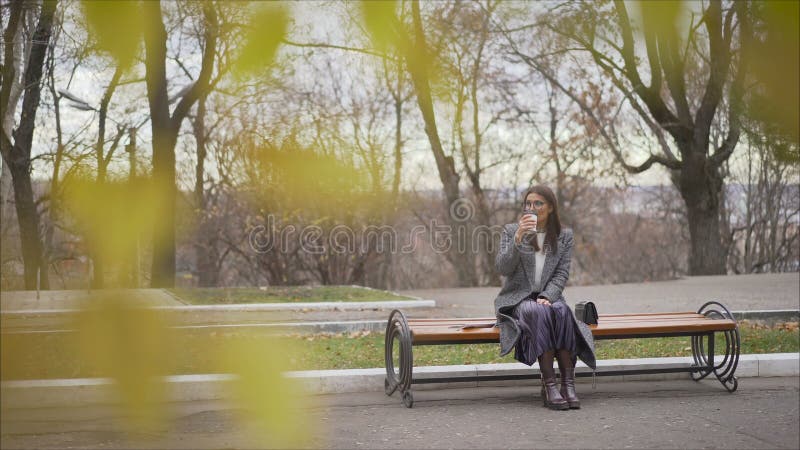 Beautiful girl in a coat sitting on a bench in a sunny park in autumn. Girl drinks warm coffee and outdoors. Portrait of