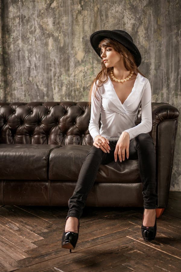 Portrait of beautiful young business woman in black hat and white shirt on leather sofa