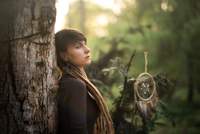 Portrait of beautiful young woman with dreadlocks in dark dress with traditional american indian amulet dreamcatcher standing back