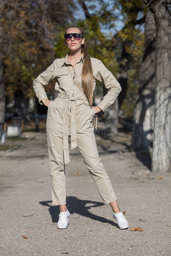 Portrait of blonde person in a jumpsuit in public Park. Attractive young woman in sunglasses and overalls similar to the uniform posing in autumn park with arms akimbo