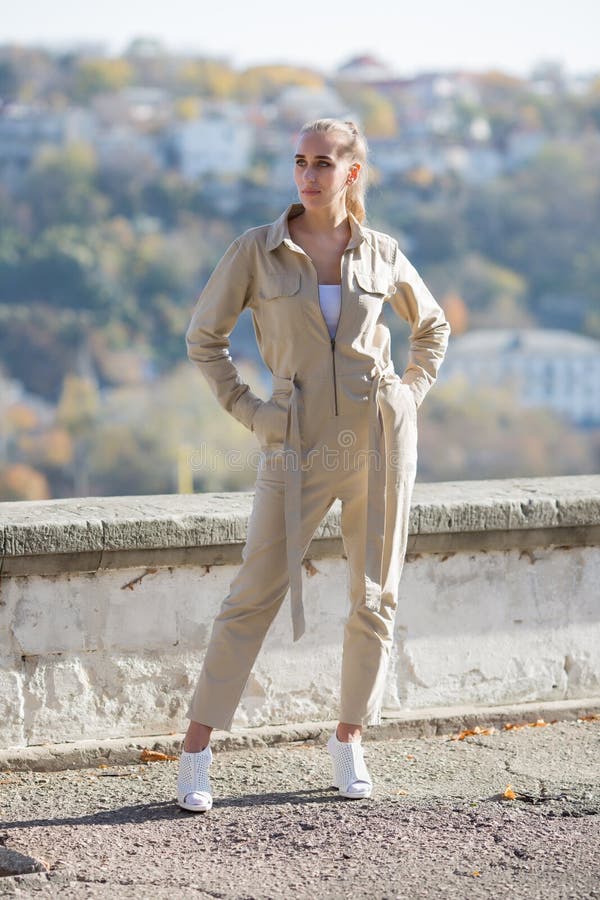 Portrait of blonde person in unbuttoned jumpsuit in public Park. Attractive young woman in sunglasses and overalls similar to the uniform posing against retaining wall park with arms akimbo