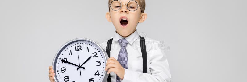 A charming boy in a white shirt, suspenders, a tie and light jeans stands on a gray background. The boy is holding a